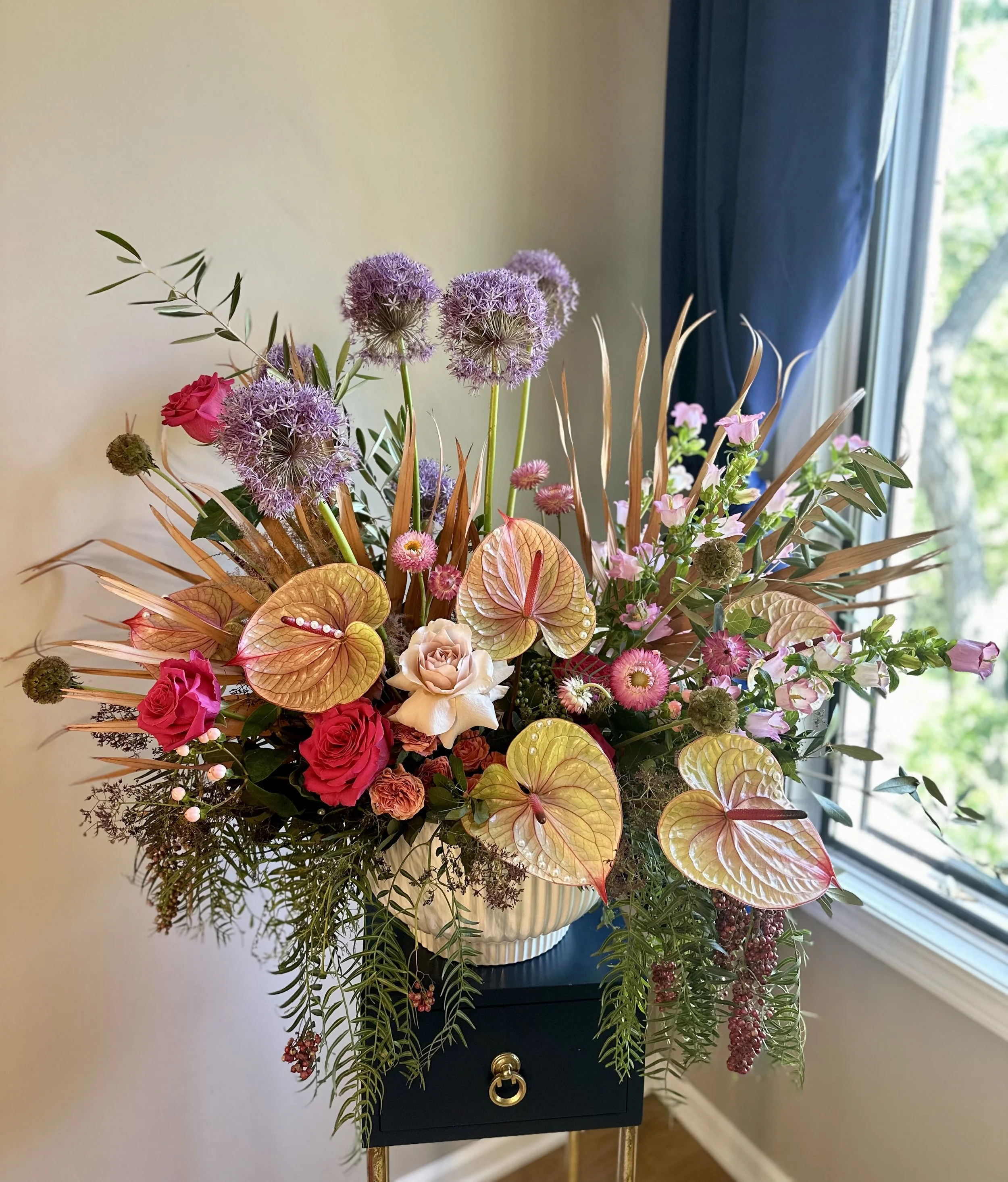 A vibrant bouquet of various colorful flowers including pink roses, purple alliums, pink and white anthuriums, and anthuriums with heart-shaped leaves, arranged in a white ceramic vase on a dark small table near a window with blue curtains.