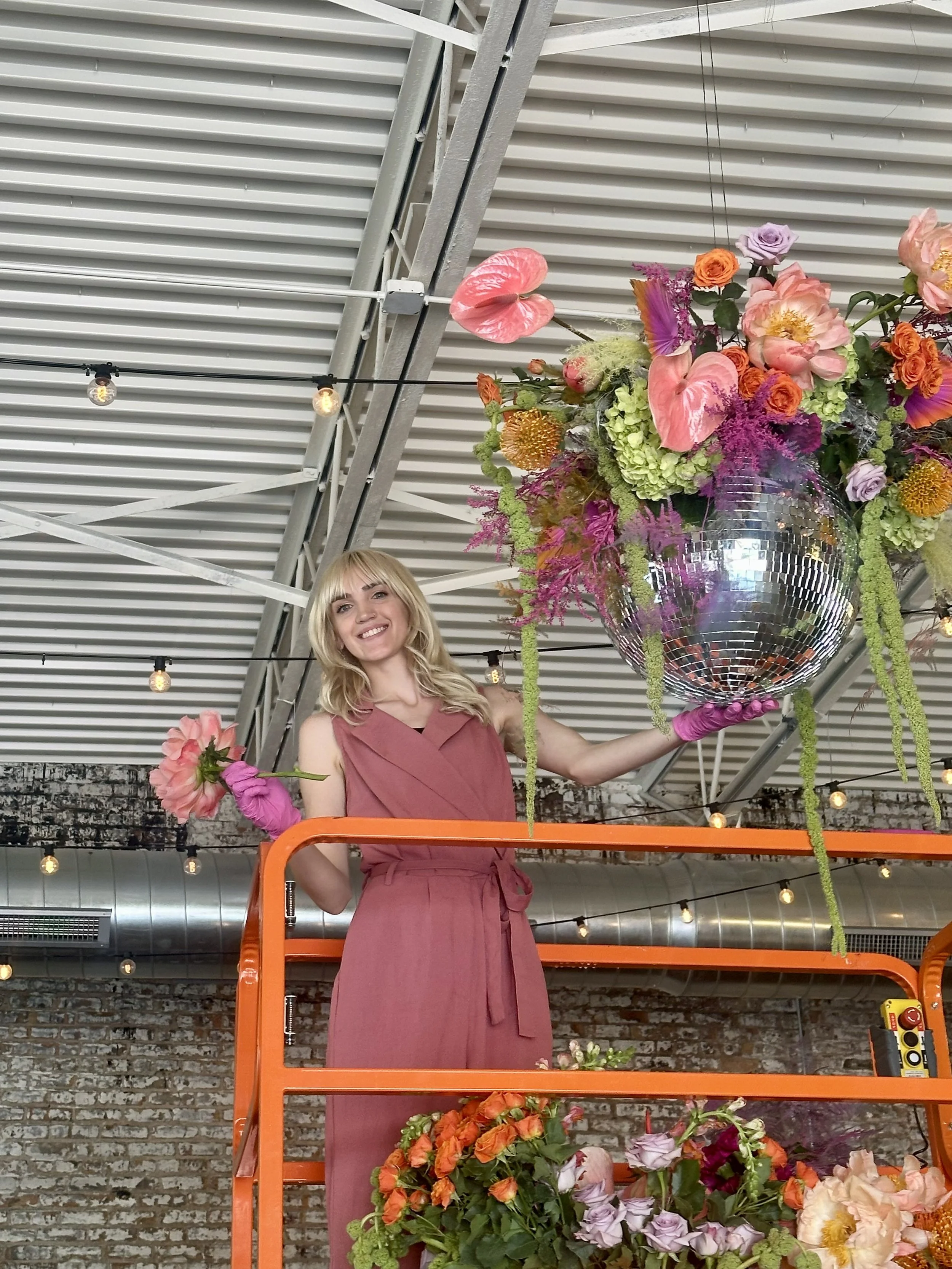 Woman in a pink dress standing on an orange lift, holding a bouquet of flowers, with large floral arrangement and disco ball overhead, in an industrial-style space with exposed brick wall and string lights.