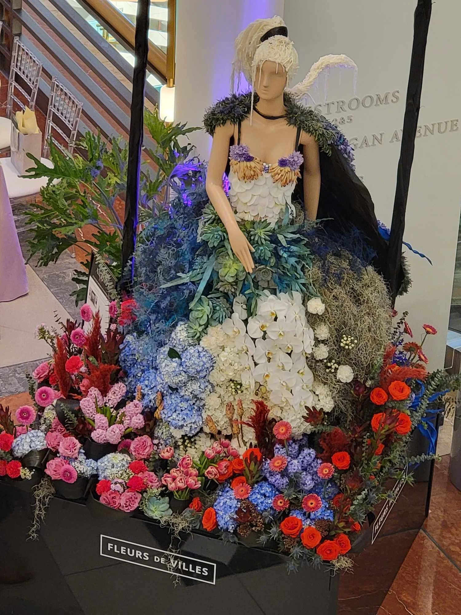 A mannequin dressed in a dress made of colorful flowers, surrounded by an arrangement of pink, red, white, purple, and blue flowers, with a black and white sign reading 'FLEURS DE VILLES' in front.