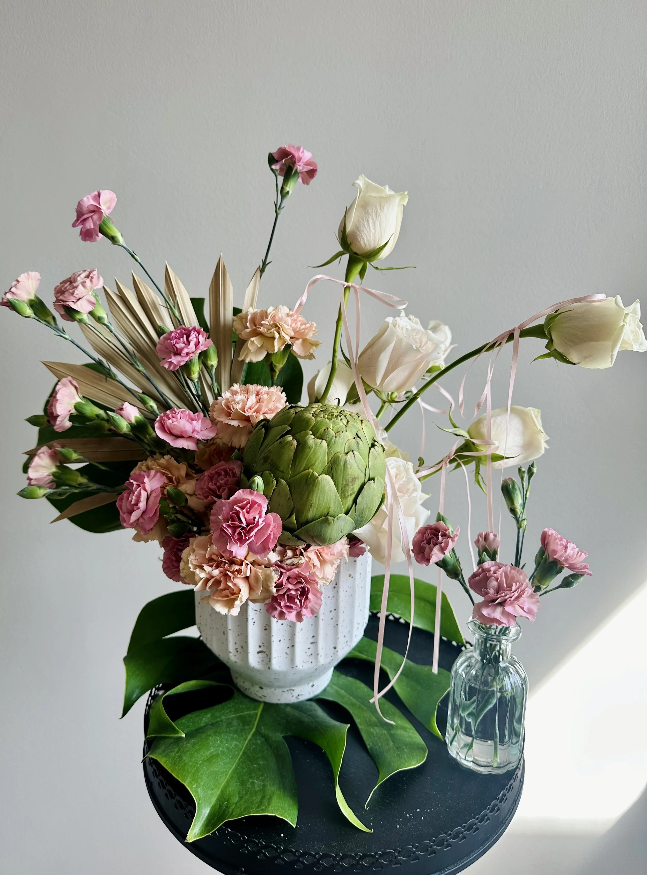 A floral arrangement with pink and cream flowers, a green artichoke, and large green leaves in a white vase, placed on a black table with a plain gray wall background.