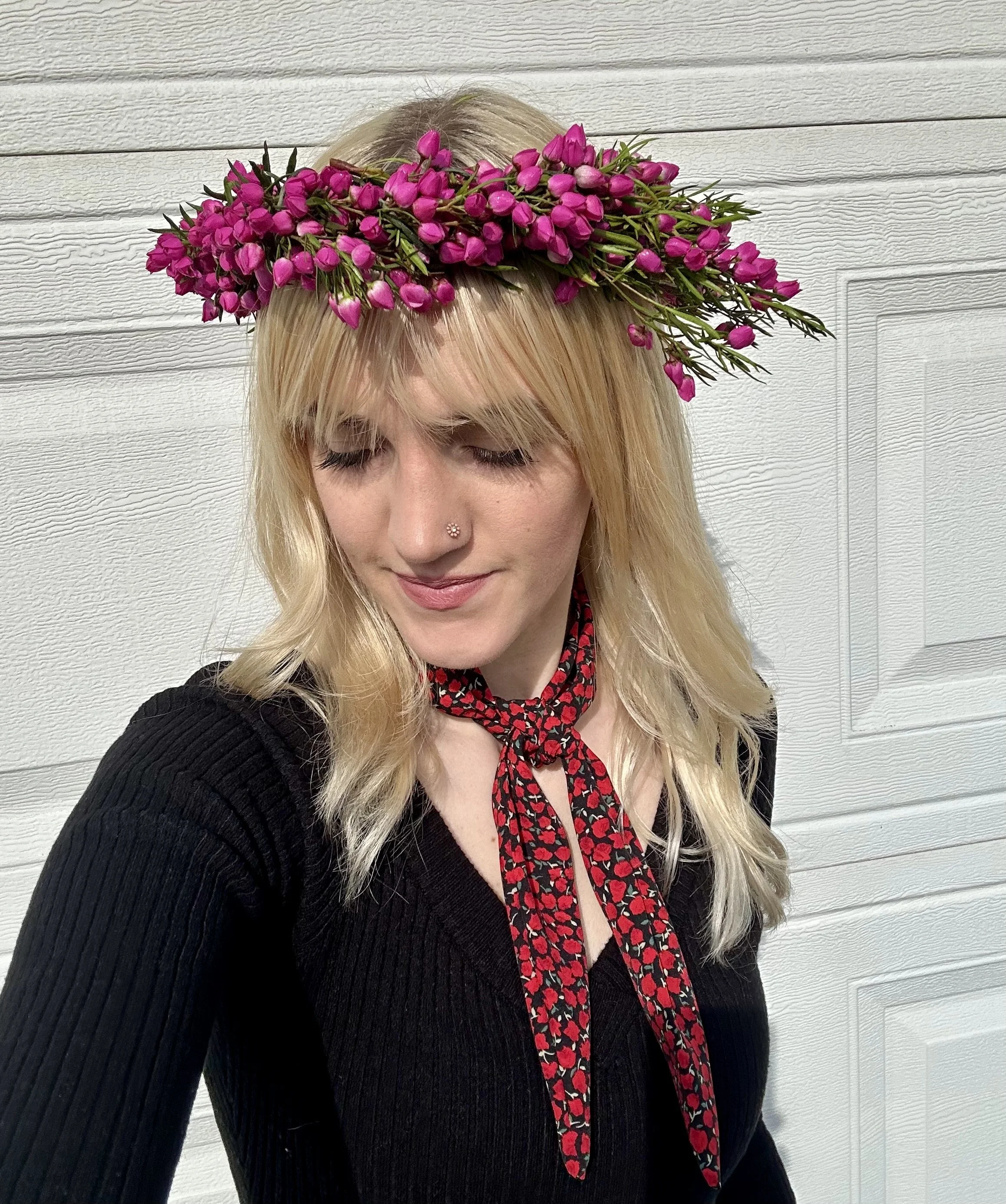 A woman with blonde hair wearing a pink flower crown, a black top, and a black and red floral scarf against a white wooden wall.