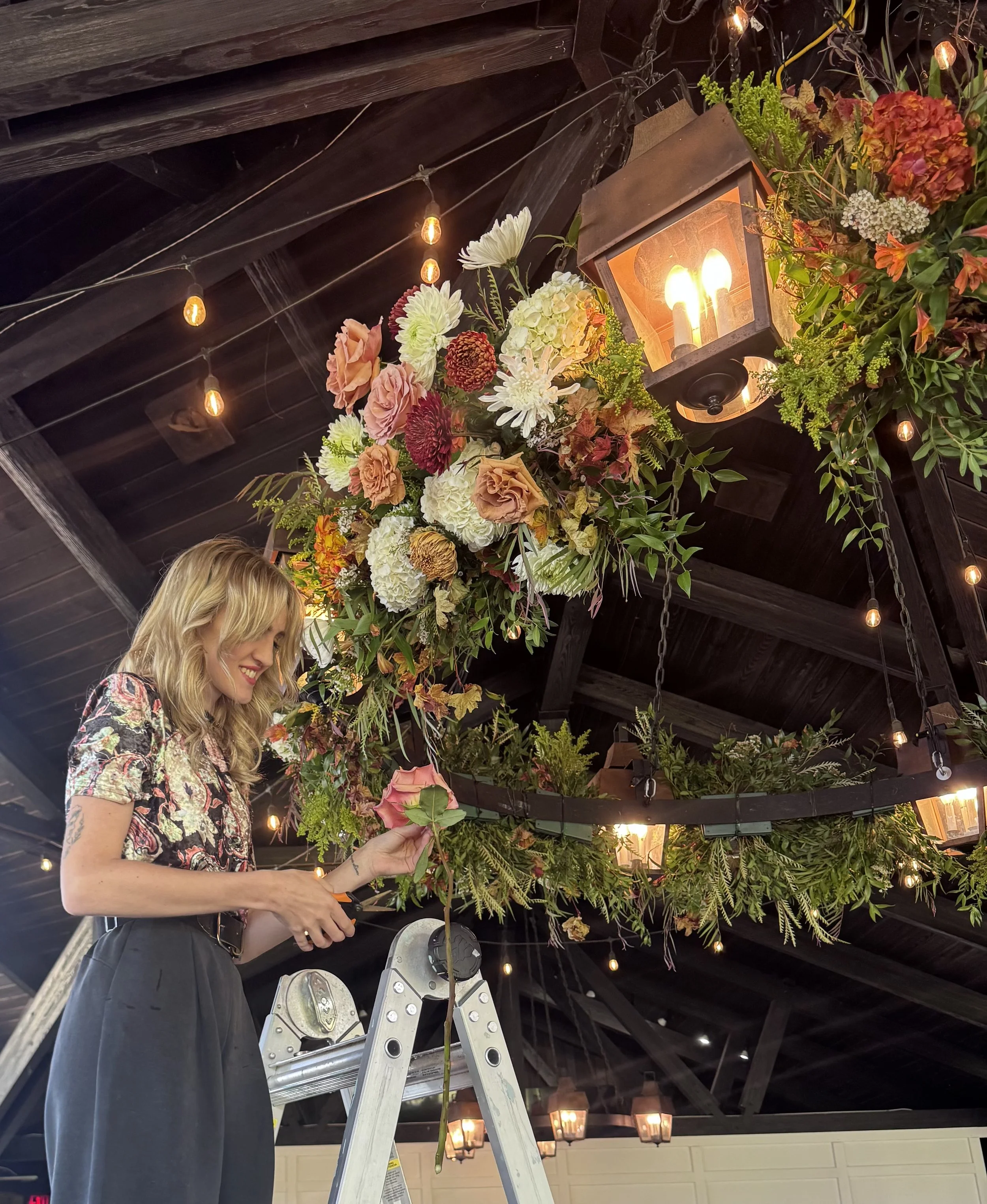A woman with blonde hair decorating a ceiling with flowers and lanterns while standing on a ladder.