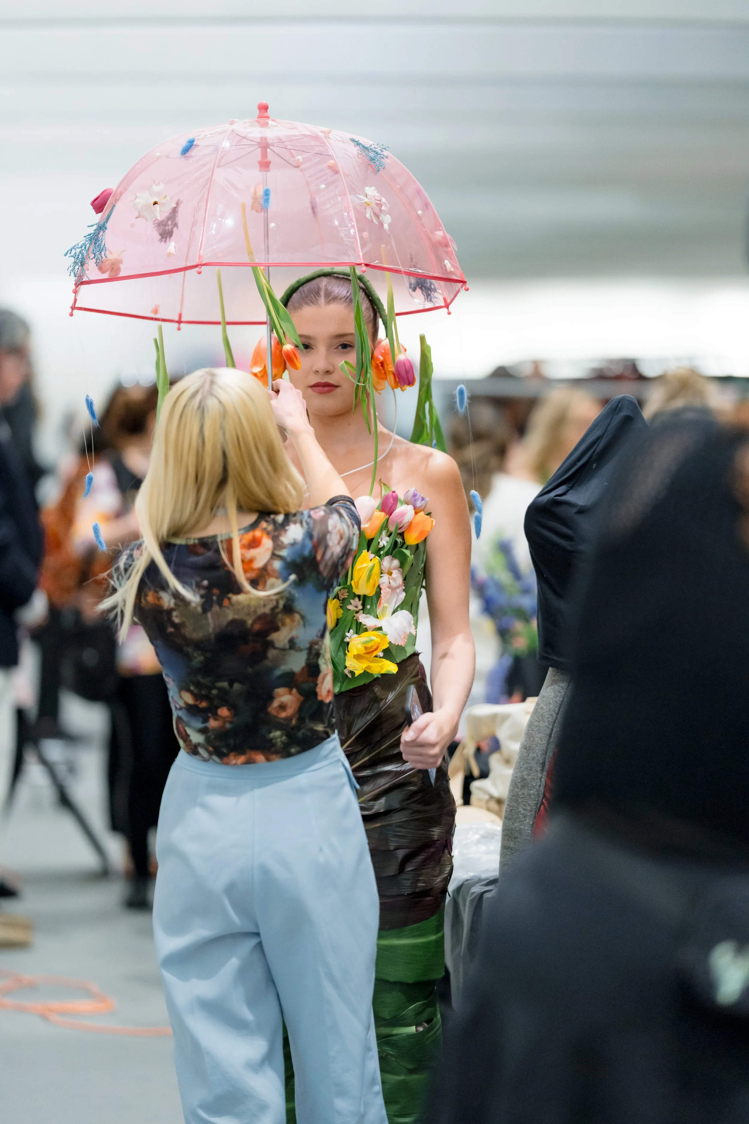 A woman wearing a floral dress is being styled with tulips on her body and an umbrella decorated with flowers and butterflies, standing in a crowded indoor space.
