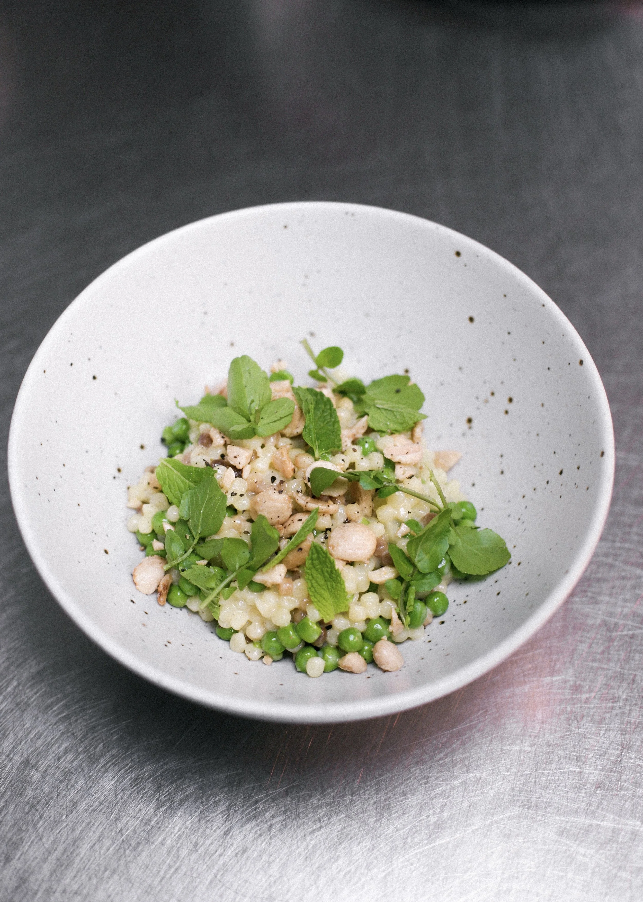 A white bowl containing cooked pearl barley salad with chopped chicken, green peas, and fresh mint leaves, set on a stainless steel surface.