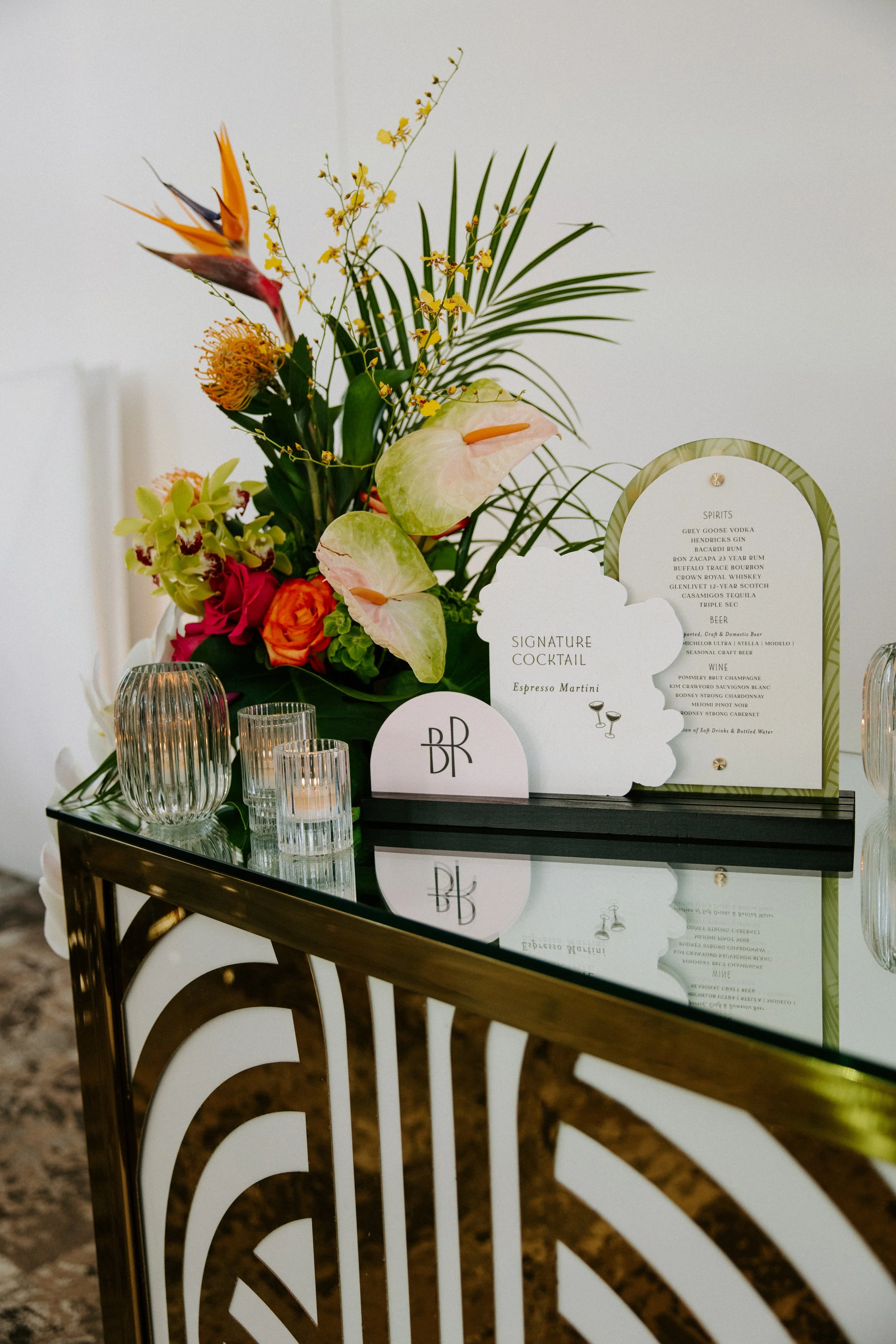 Elegant bar station with a colorful flower arrangement, glassware, and cocktail and beverage menus on a reflective surface.