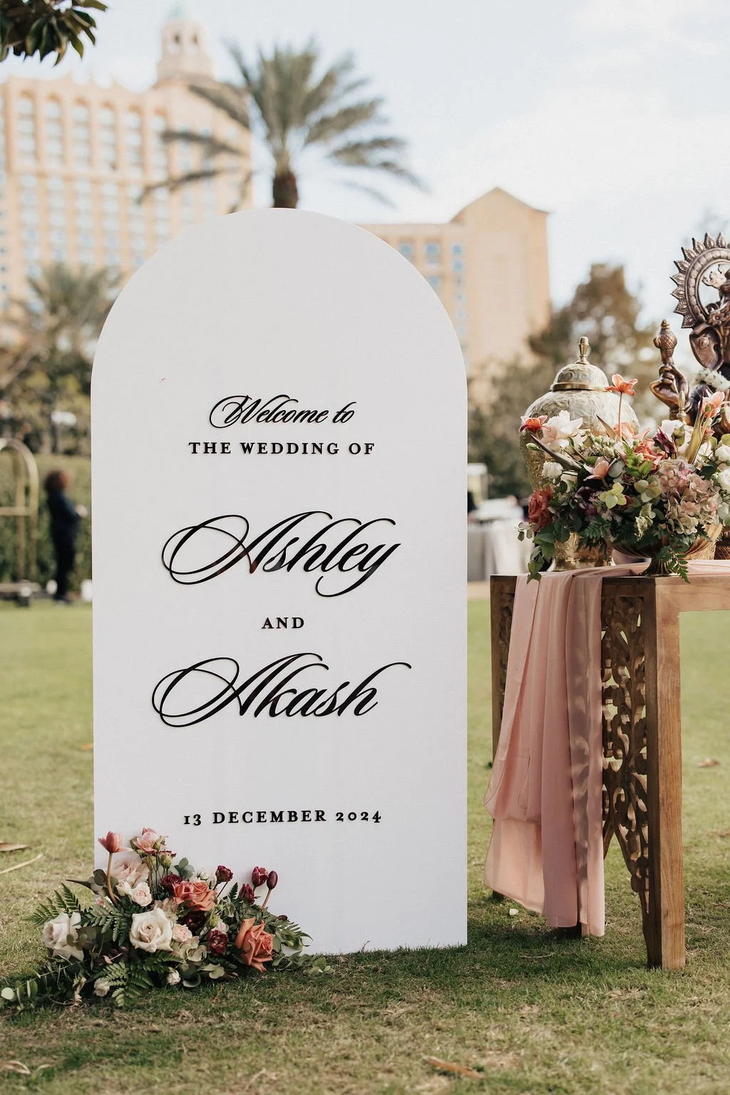 Wedding welcome sign with floral arrangements on a grassy outdoor area, with tall buildings and palm trees in the background. The sign reads: "Welcome to the wedding of Ashley and Nash, 13 December 2024."