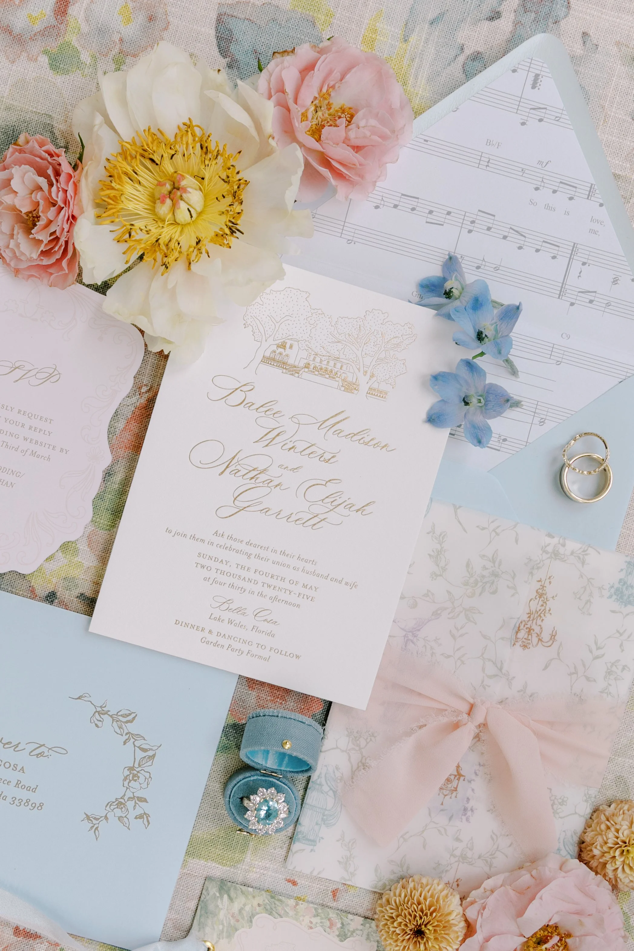 Wedding invitation with floral decorations including pink and white flowers, blue flowers, and gold rings, on a pastel-colored background with sheet music and other stationery.
