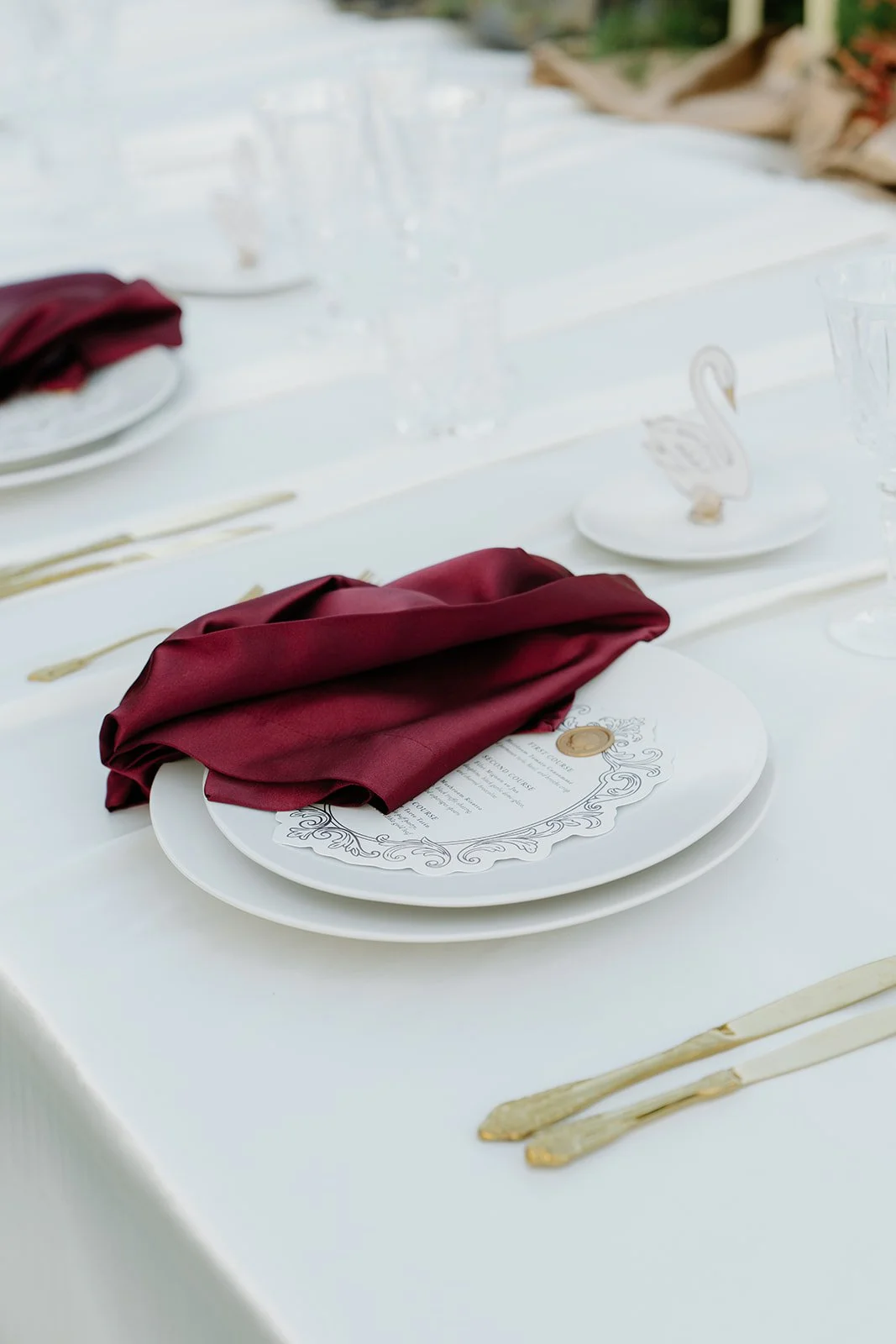 Elegant table setting with white plates, gold-colored cutlery, a dark red napkin, and a menu with decorative border and seal, all on a white tablecloth.