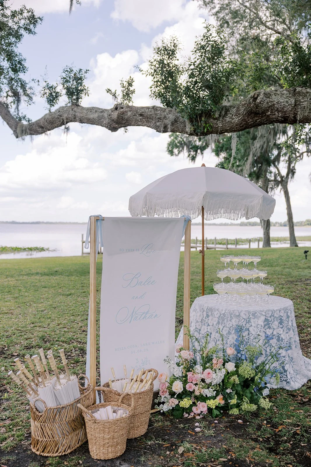 Wedding setup with a white umbrella, a sign for the couple Sallee and Nathan, a table with glasses, and a floral arrangement near a lake.