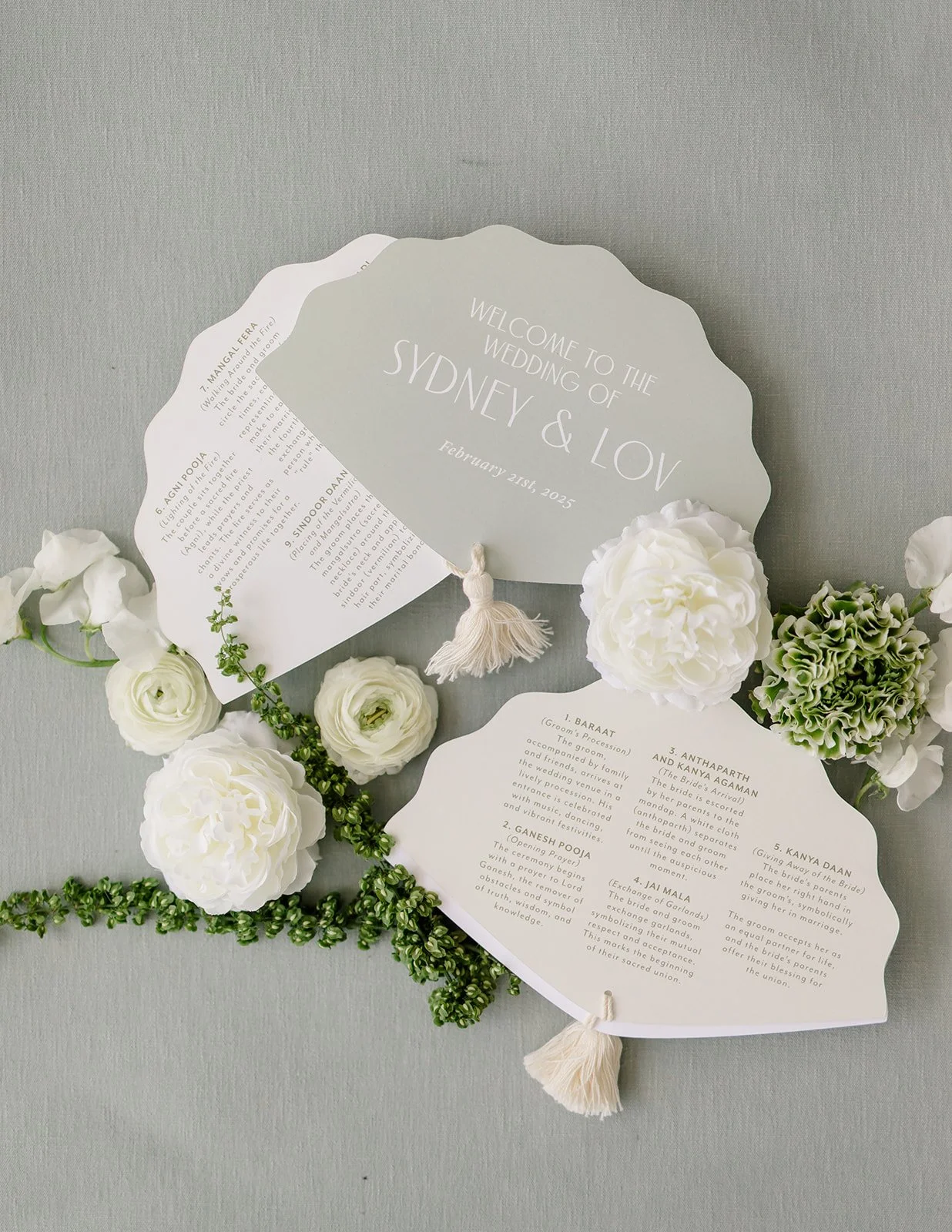 Wedding program and welcome sign surrounded by white and green flowers on a light gray surface.