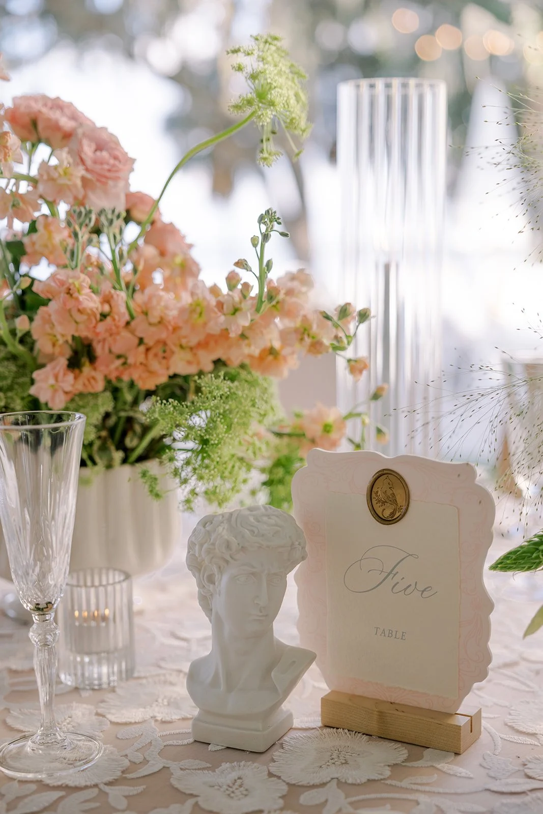 Elegant table centerpiece with pink flowers, a small white bust sculpture, a clear glass vase, and table number sign on lace tablecloth.