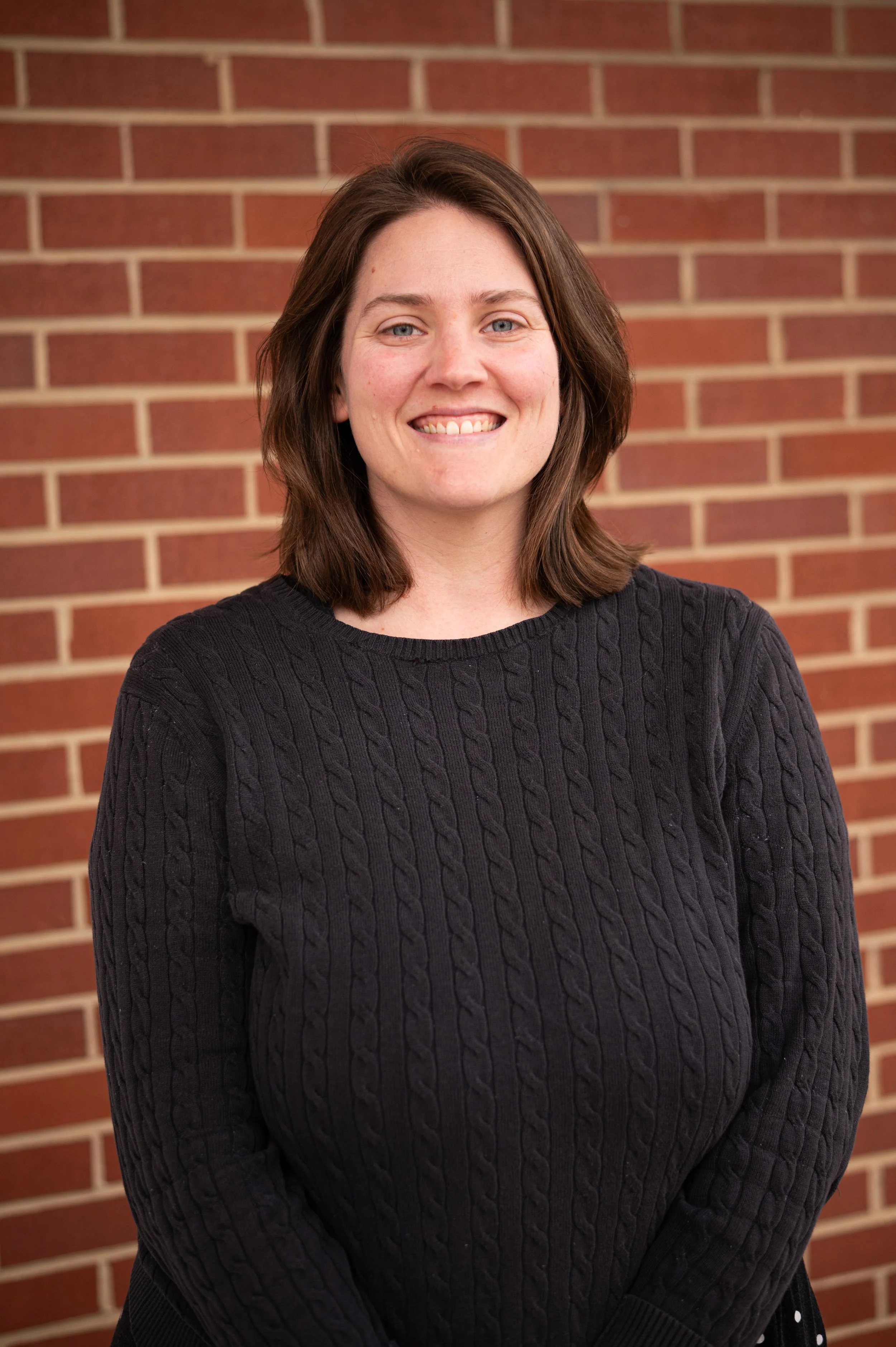 A woman with long brown hair and a sleeveless light-colored blouse smiling in front of a red brick wall.