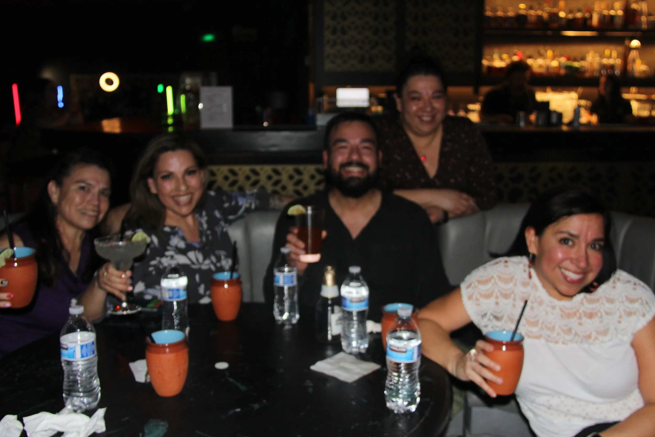 Group of people smiling and holding drinks at a bar table with background lighting.