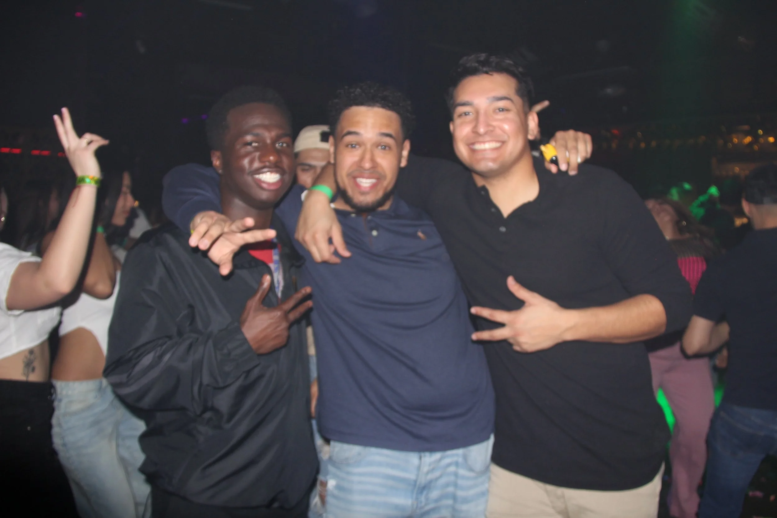 Three men smiling and posing for a photo in a club setting, surrounded by people dancing.