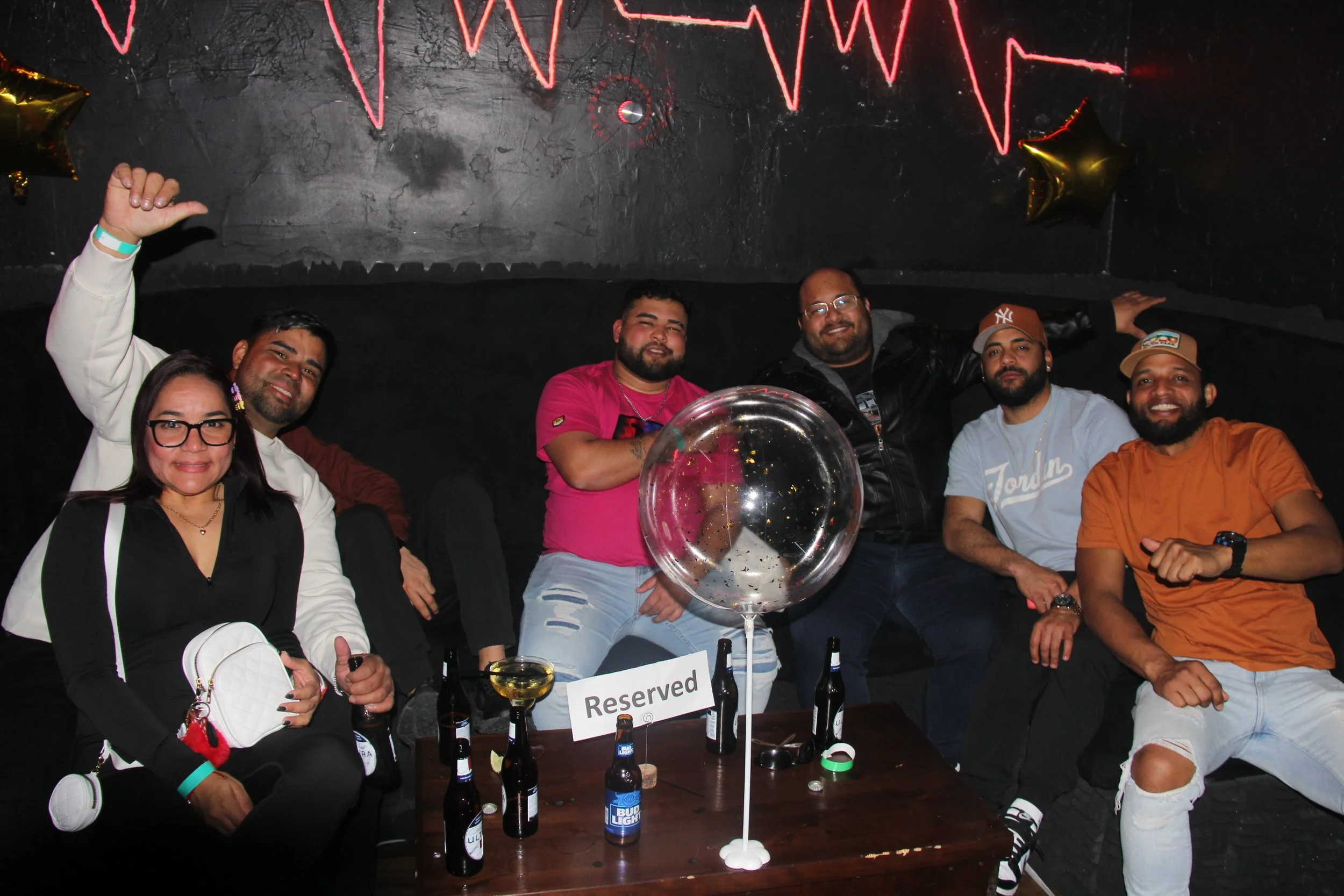 Group of six people sitting in a lounge with a reserved table, beer bottles, and a large transparent balloon; black wall with neon light design in the background.