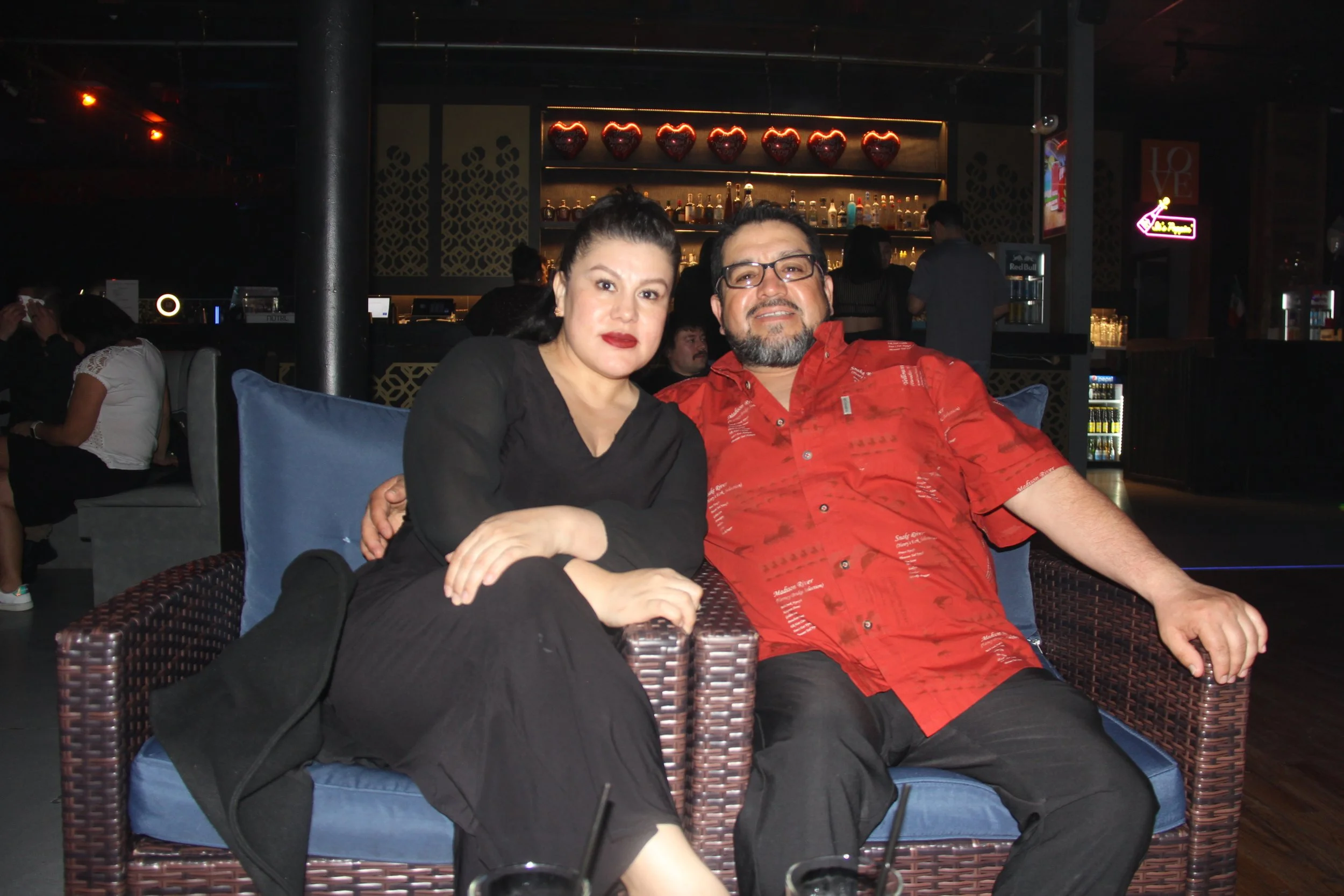 A couple sitting on a wicker chair at a bar, with glasses on the table in front of them and heart-shaped decorations in the background.
