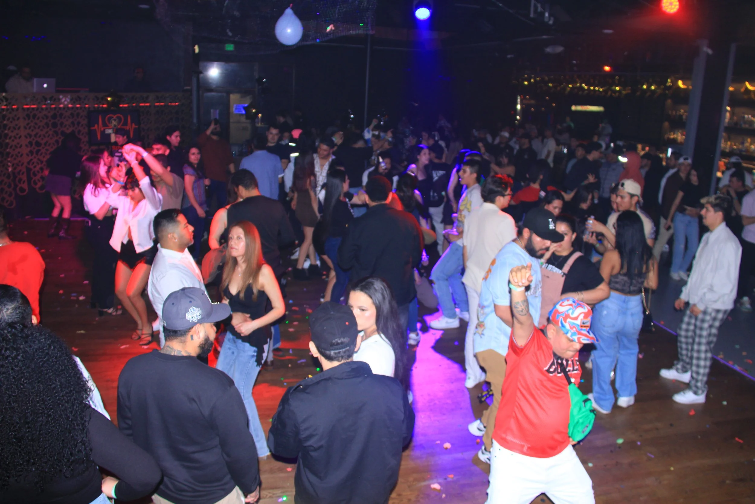 People dancing in a crowded nightclub with colorful lighting and a DJ booth in the background.