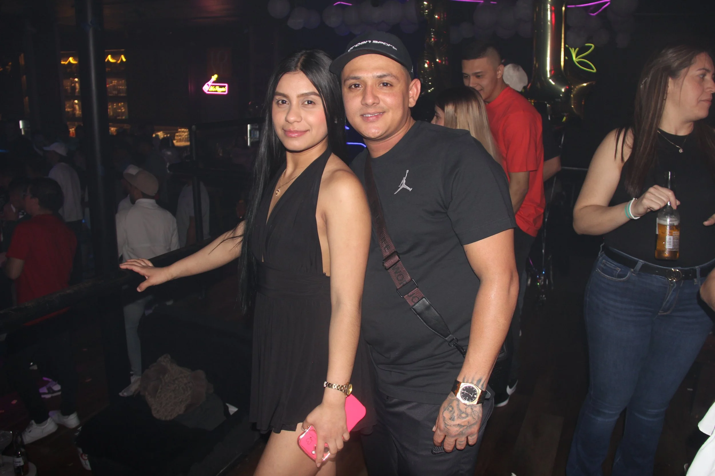 People enjoying a night out at a club, with a woman in a black dress holding a phone and a man in a black cap and t-shirt posing together. Other patrons are in the background with drinks.