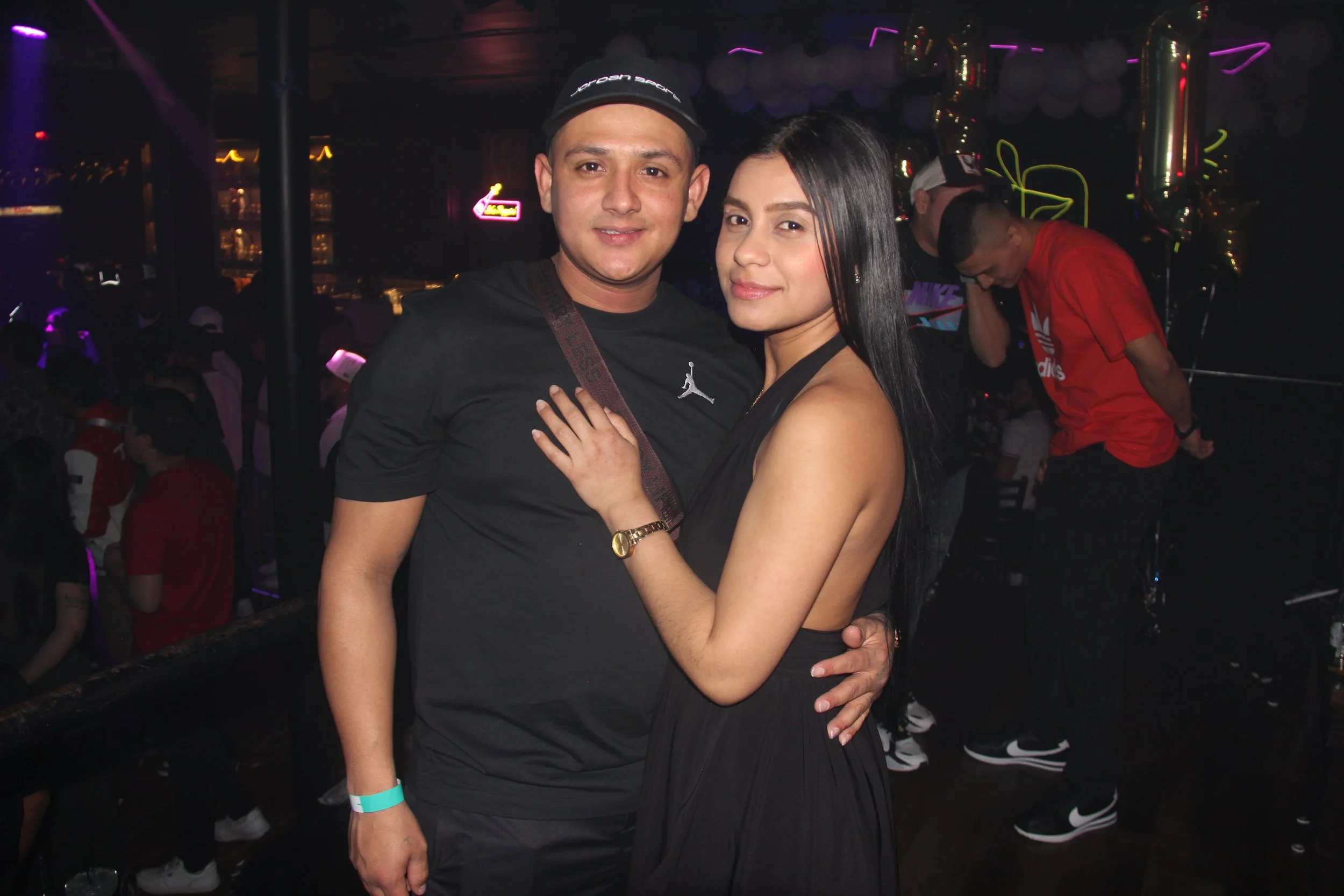 A couple posing in a dimly lit club with people in the background, balloons, and neon lights.