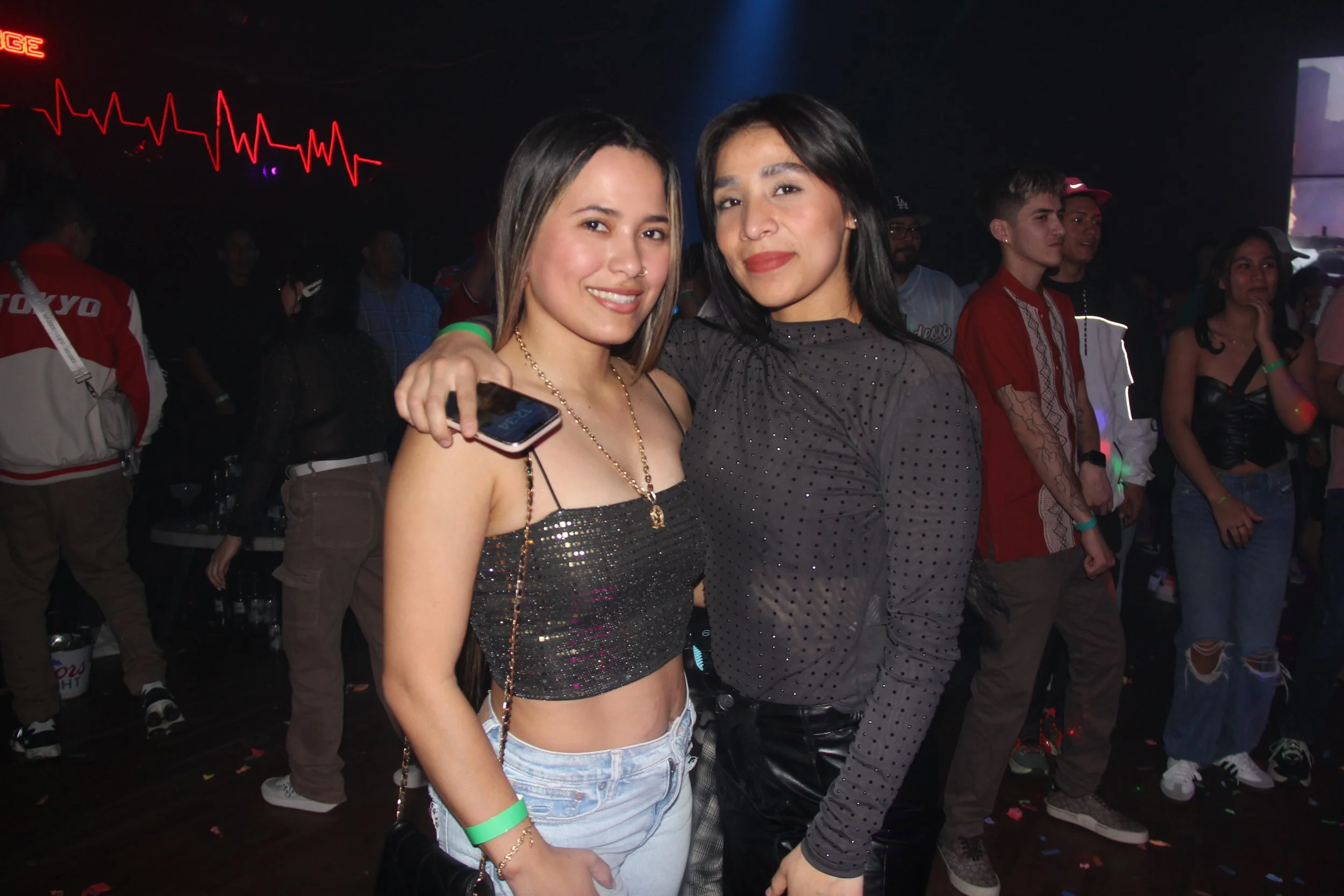 Two women posing for a photo at a nightclub, with a crowd and neon sign in the background. One holds a phone and wears a sparkly top, while the other wears a dotted black top.