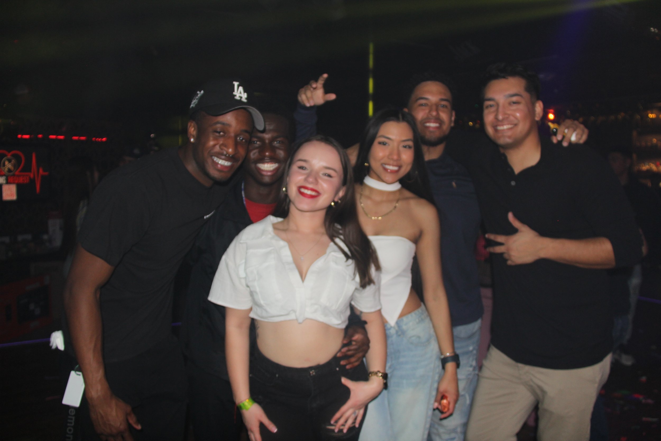 Group of friends posing together in a dimly lit club or party setting, smiling and appearing to have a good time.