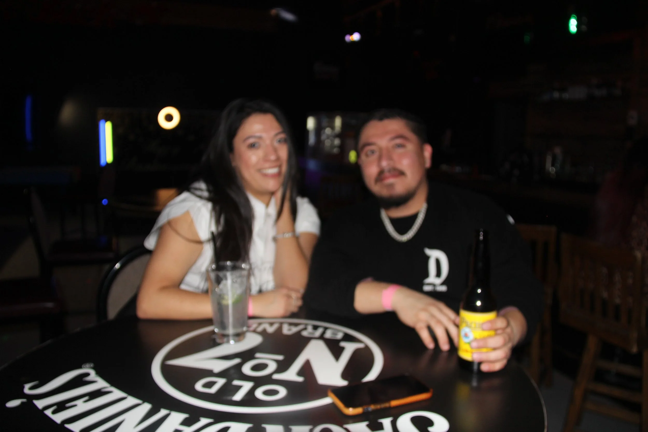 A man and woman sitting at a round table in a dimly lit bar, smiling at the camera, with drinks and a phone on the table with a Jack Daniel's logo.