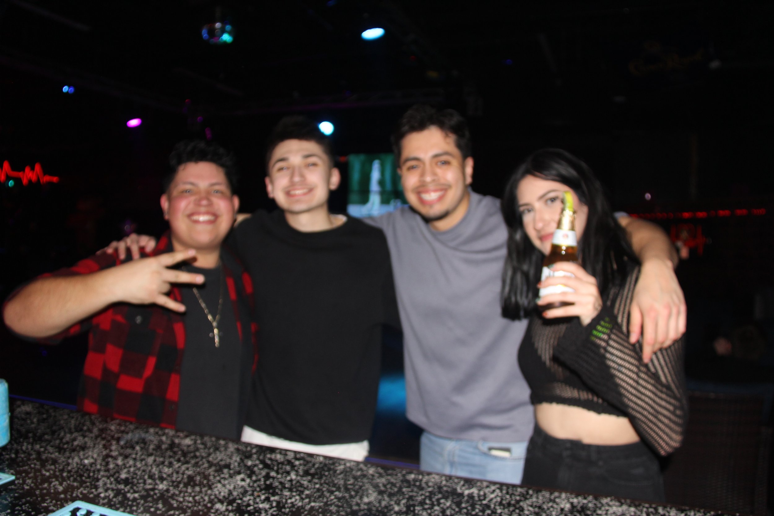 Group of four friends at a nightclub, three men and one woman, smiling and posing by a bar counter. The woman is holding a beverage bottle with a lime slice.