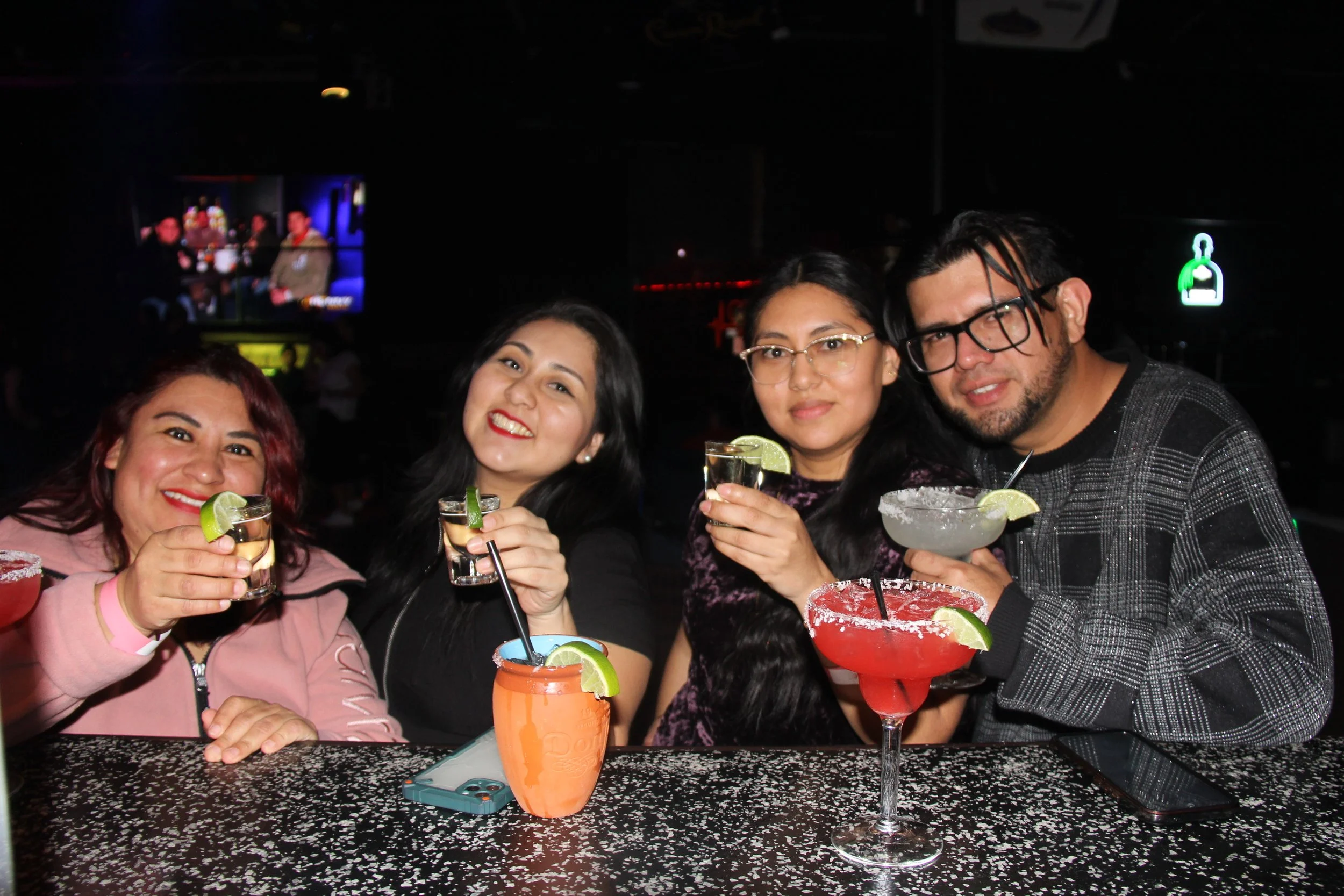 Group of four people at a bar holding various cocktails with lime garnishes, smiling at the camera.