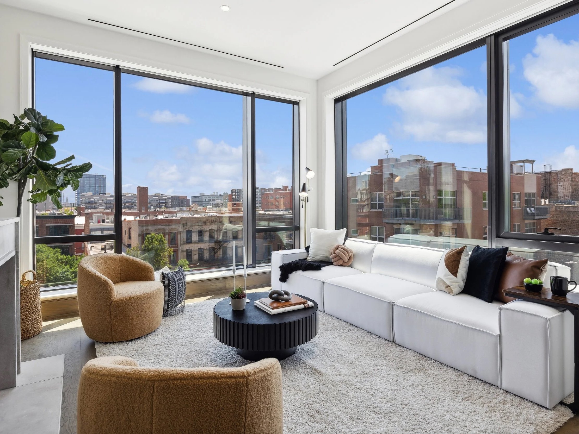 Modern living room with large windows, white sofa, tan armchairs, black coffee table, rug, and city view outside.