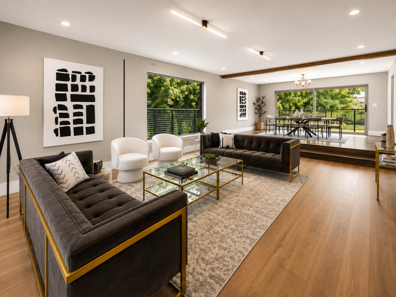 Modern living room with black and white abstract art, two white armchairs, dark couches with throw pillows, glass and gold coffee table, rug, large windows, and a dining area on a slightly elevated platform.