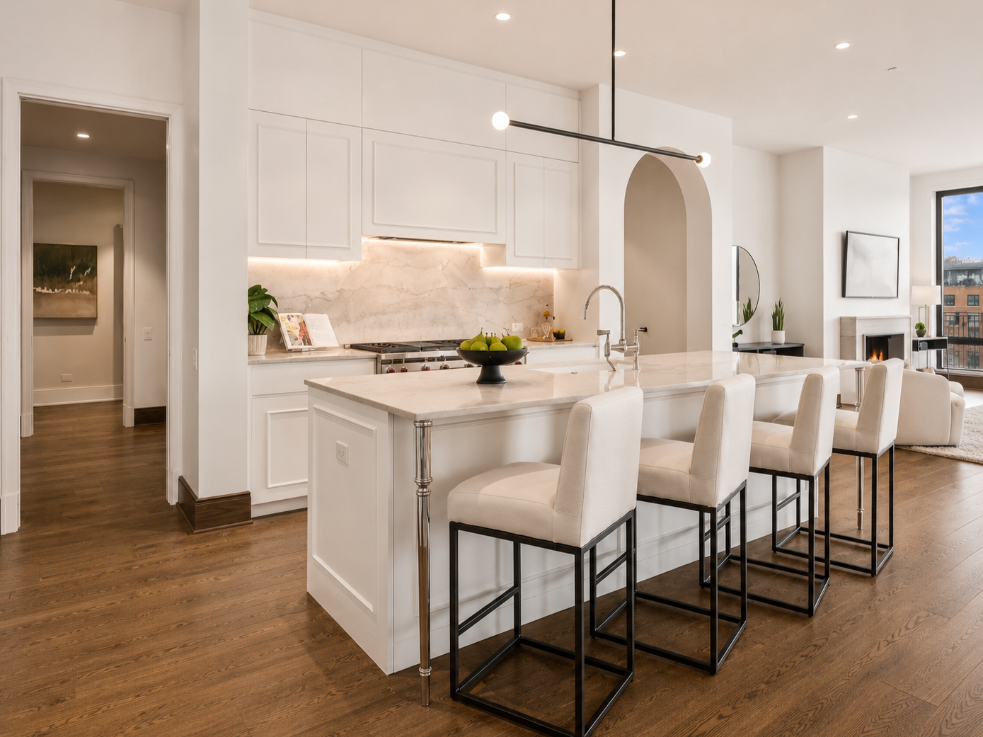 Modern kitchen with white cabinets, marble countertops, and a kitchen island with cream-colored stools. The space is bright with natural light and has a living area with a fireplace and large window.