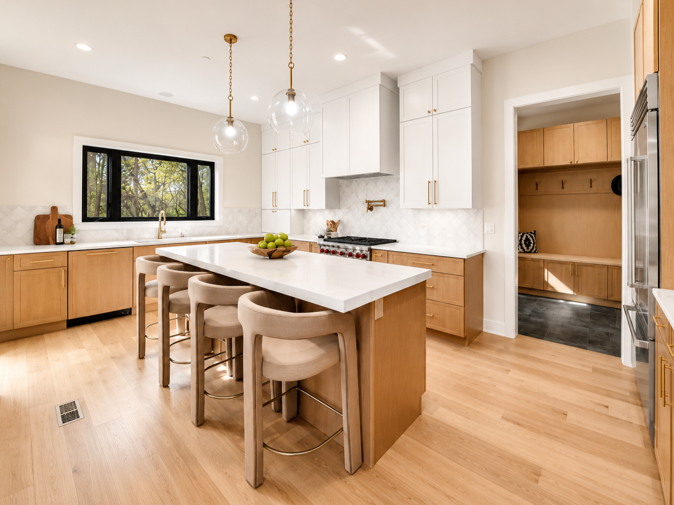 Modern kitchen with light wood cabinets, white countertops, a central island with beige bar stools, a black window, and a separate pantry area with built-in seating.