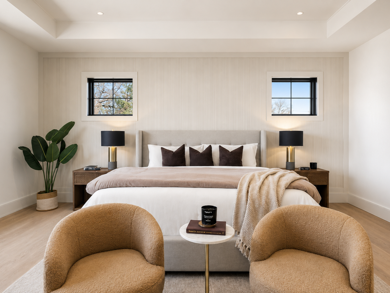 Modern bedroom with a king-size bed, two symmetrical nightstands with black lamps, a potted plant in the corner, two small windows, beige chairs in front of the bed, and neutral-colored decor.