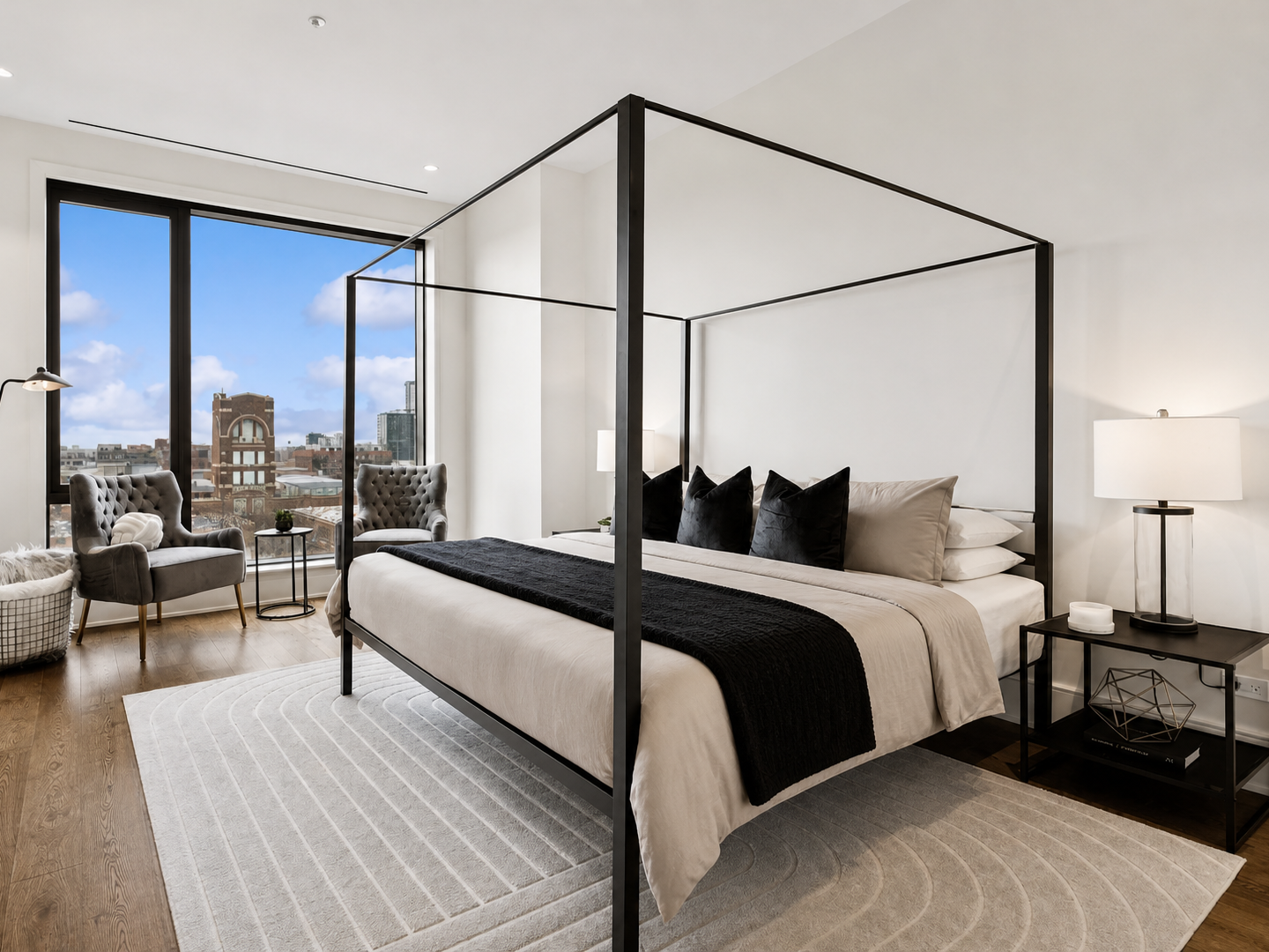 Modern bedroom with a four-poster bed, black pillows, beige bedding, and large windows showing a city view. The room features a white area rug, two gray armchairs near the window, a black side table with a lamp and decorative items, and a basket of b