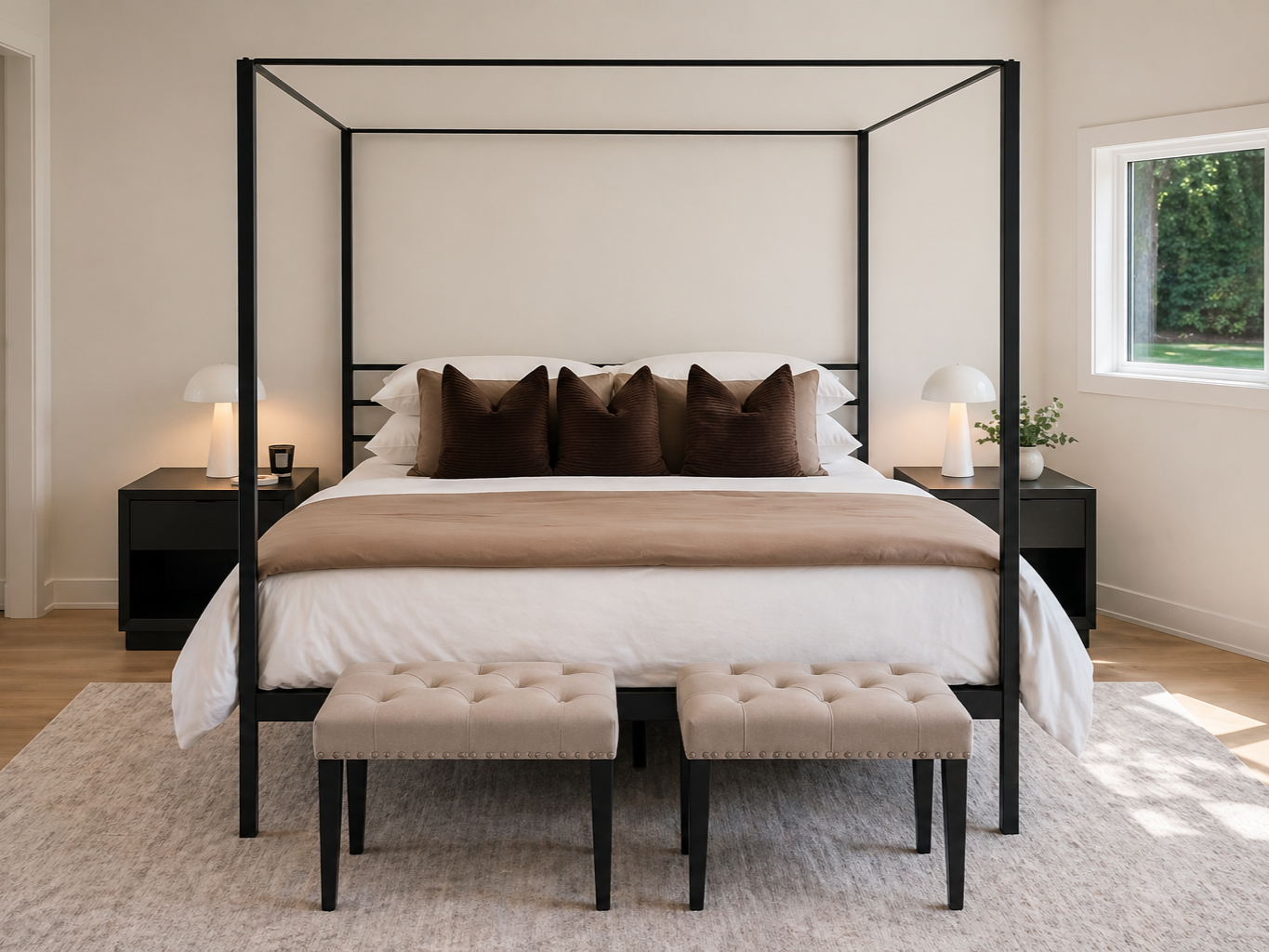 Modern bedroom with a black canopy bed, beige bedding, brown pillows, two black nightstands with white lamps, a beige tufted bench at the foot of the bed, a window with greenery outside, and a neutral-toned rug.