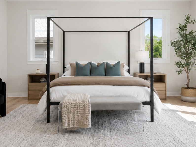 A bedroom with a black four-poster bed, white walls, two windows, wooden nightstands with lamps, a gray bench at the foot of the bed, and a potted plant in the corner.