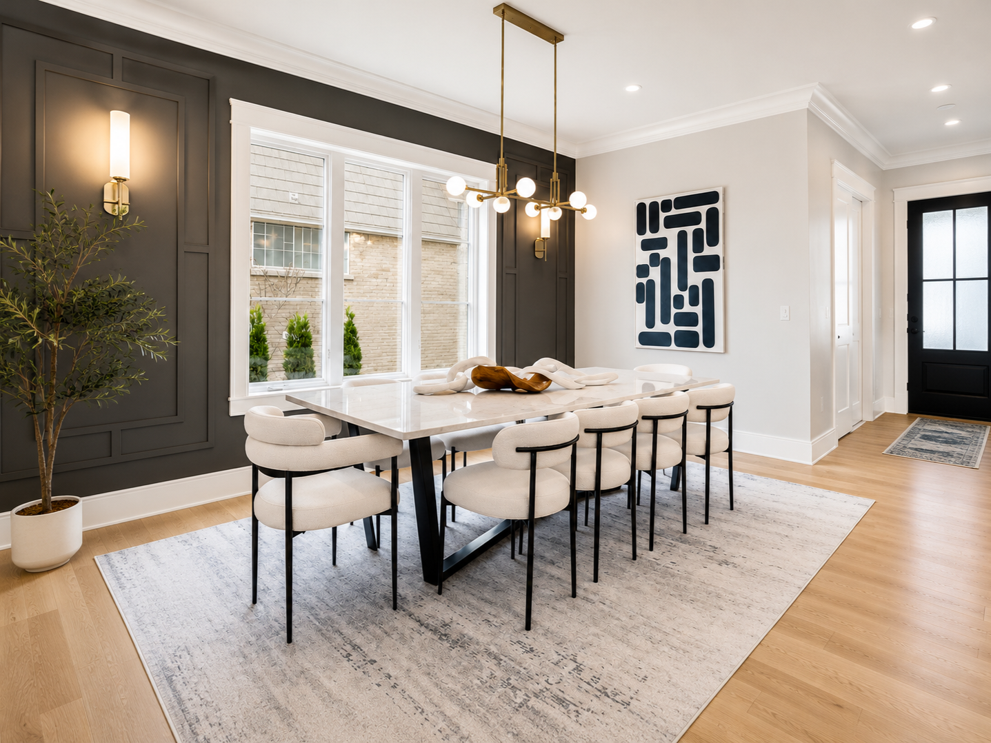 Modern dining room with white marble table, eight beige upholstered chairs with black legs, black and gold chandelier, dark gray paneled wall, large window with blinds, abstract black and white artwork, potted plant, light wood flooring, area rug, bl
