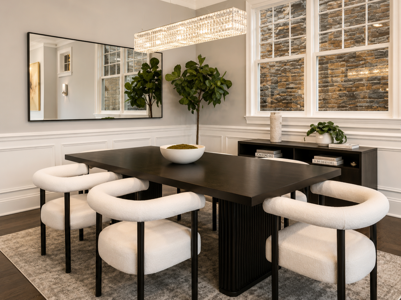 Modern dining room with a black table, six white upholstered chairs, a large mirror, and decorative items including a potted plant, a ceramic vase, and some books. The room has white walls with wainscoting, large windows with brick exterior, and a co