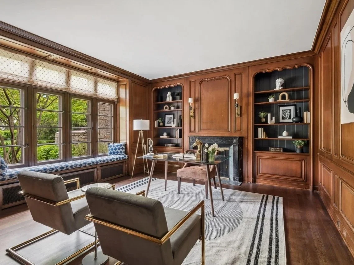A wood-paneled living room with large windows, a window seat with patterned cushions, two modern armchairs with gold accents, a desk, a fireplace with a black mantel, built-in bookshelves filled with decorative objects, books, and framed pictures, a 