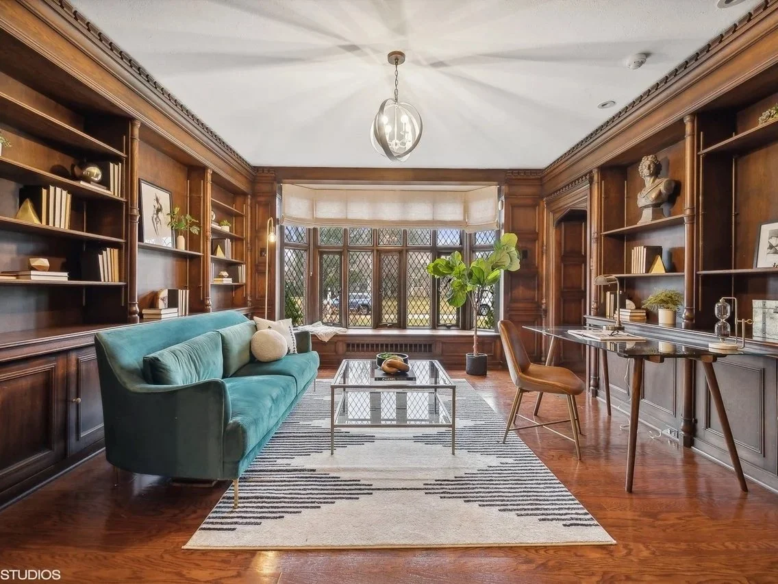 A home office or study with wood-paneled walls, built-in bookshelves filled with books and decorative items, a teal sofa, a glass-top coffee table, a wooden desk with a chair, and a large window with diamond-pane leaded glass letting in natural light