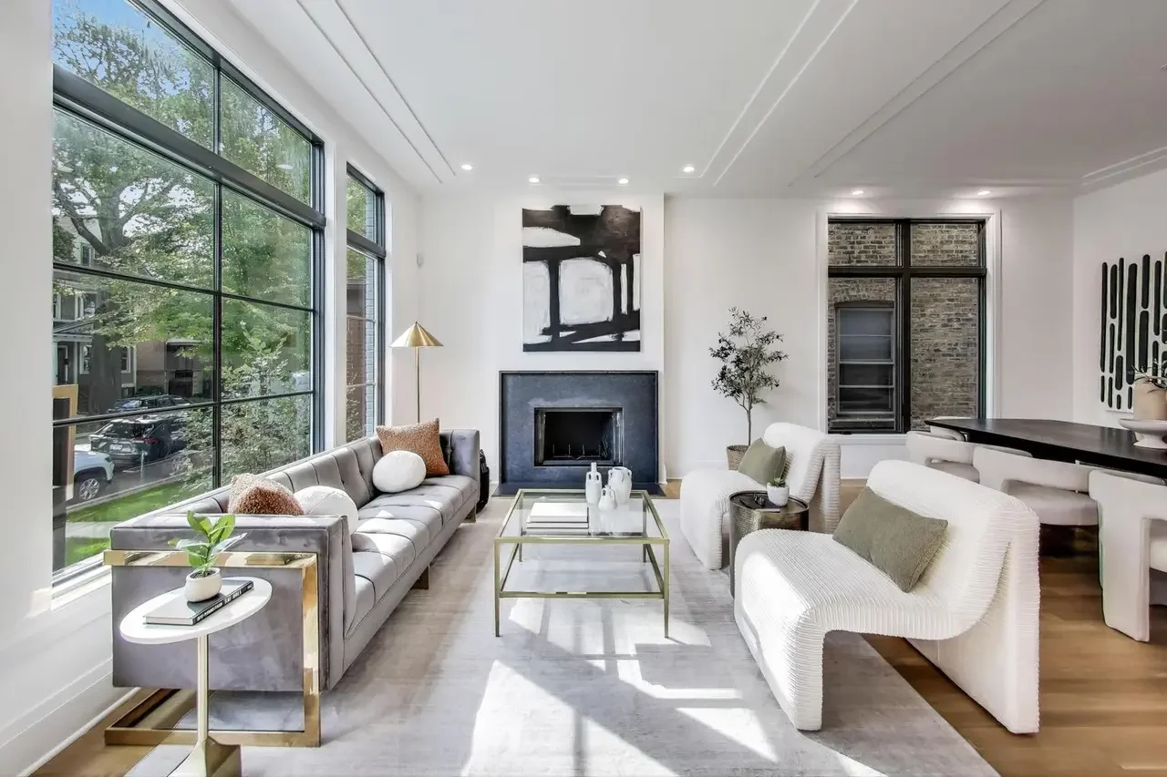 Modern living room with large windows, a gray sofa, white armchairs, a fireplace, abstract painting, and minimal decor.