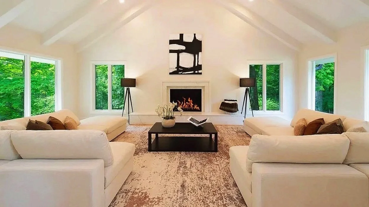 Living room with white sofas, black coffee table, fireplace, and large windows showing green trees outside.