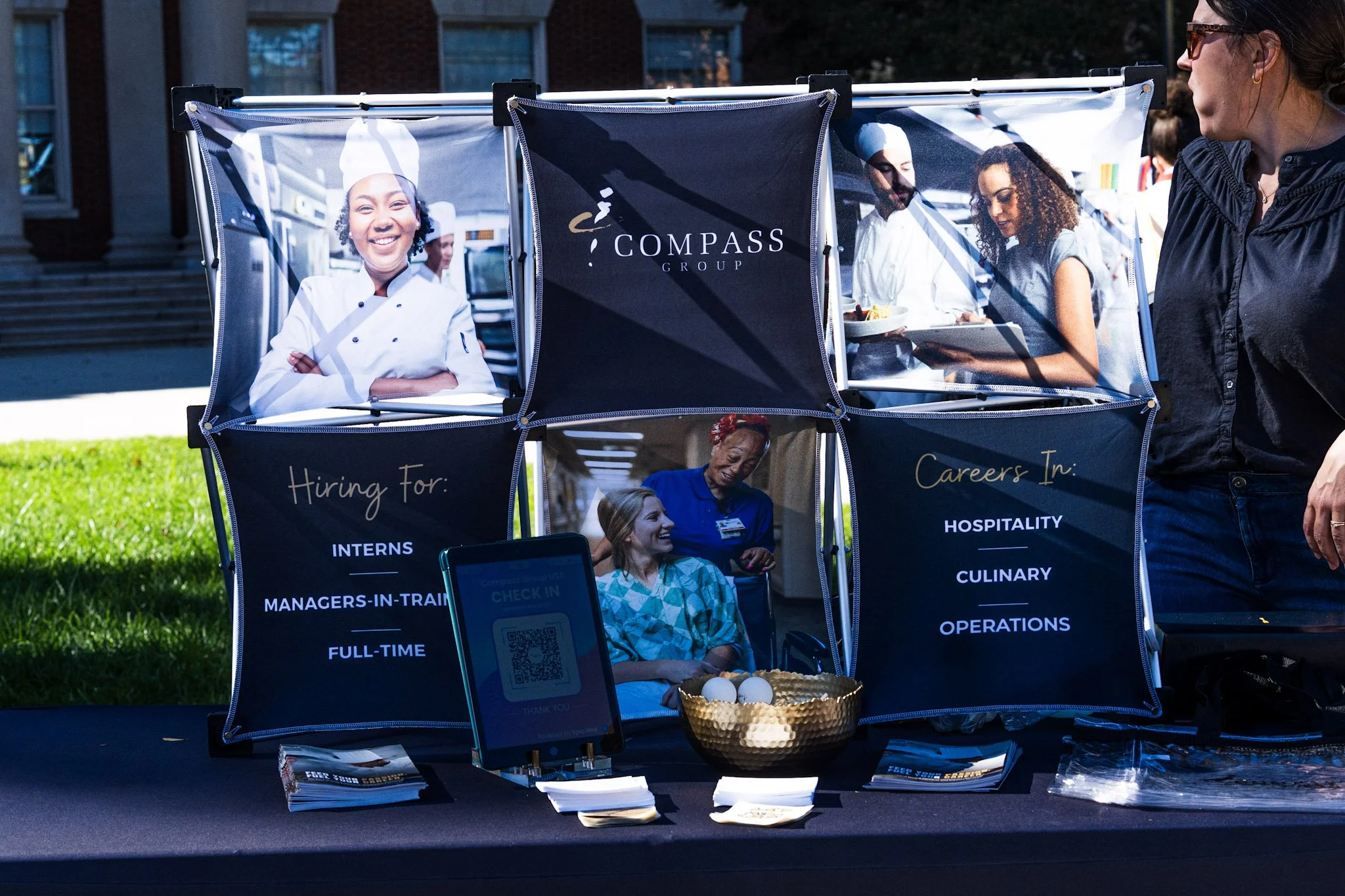 A booth at an outdoor event with posters of diverse chefs and hospital staff. The booth displays information about job opportunities in hospitality, culinary, and operations, with brochures, a QR code sign-in, and a bowl of eggs on the table.