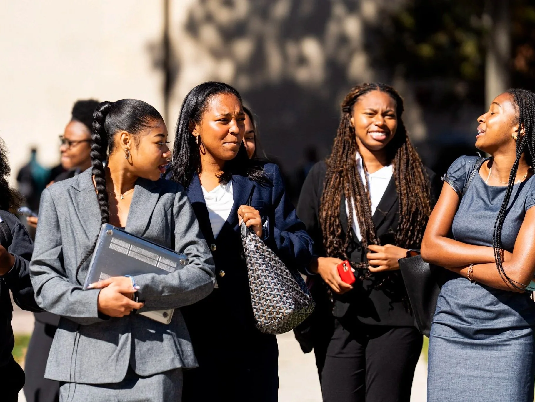 Group of professional women engaged in a conversation outdoors, dressed in business attire, some holding folders and phones.