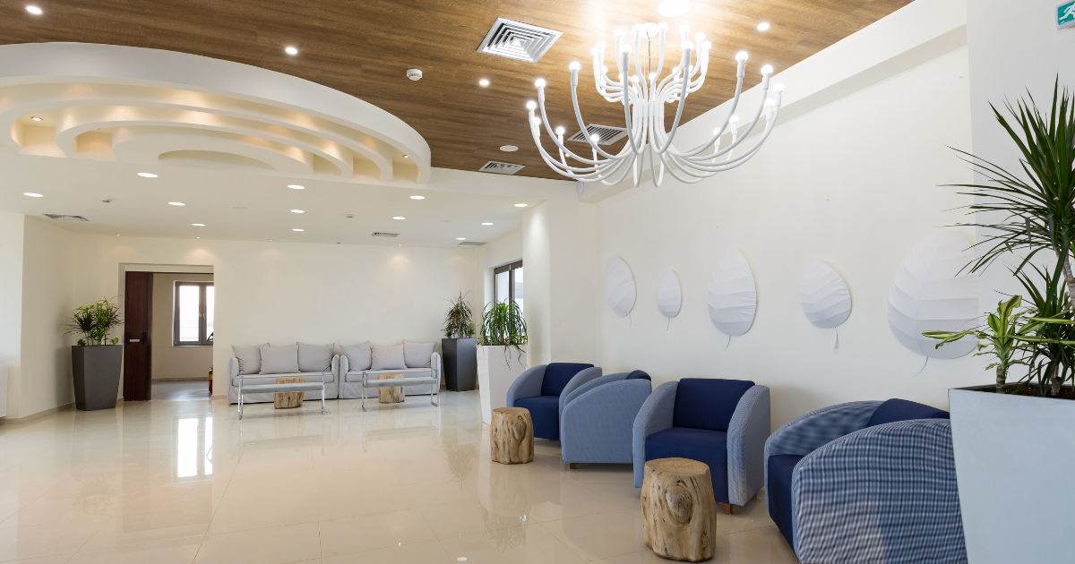 Modern waiting room with a large chandelier, blue armchairs, gray sofas, wooden stools, potted plants, and decorative wall accents.