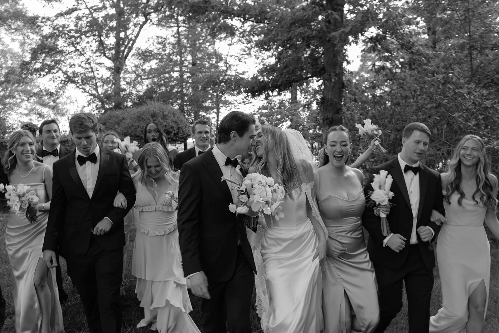 Black-and-white candid of the wedding party walking while the bride and groom sneak a kiss in the middle of the group