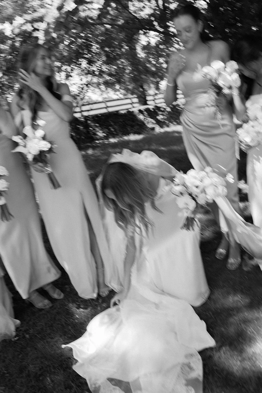 Black and white documentary wedding photograph capturing the bride adjusting her train during formal portraits with her bridesmaids.