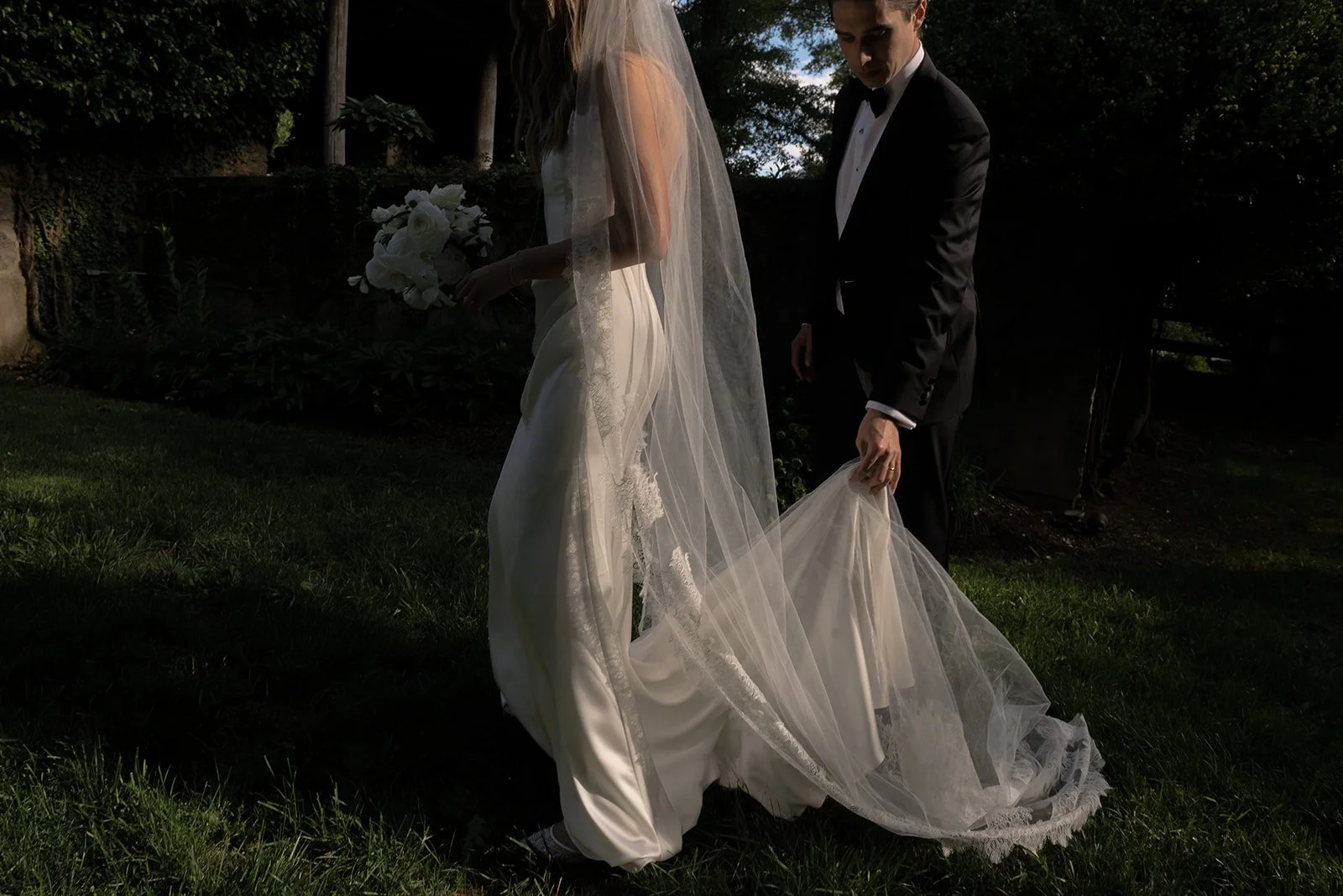 Bride and groom standing together beneath trees with the bride’s long veil draped through the grass at Goodstone Inn.
