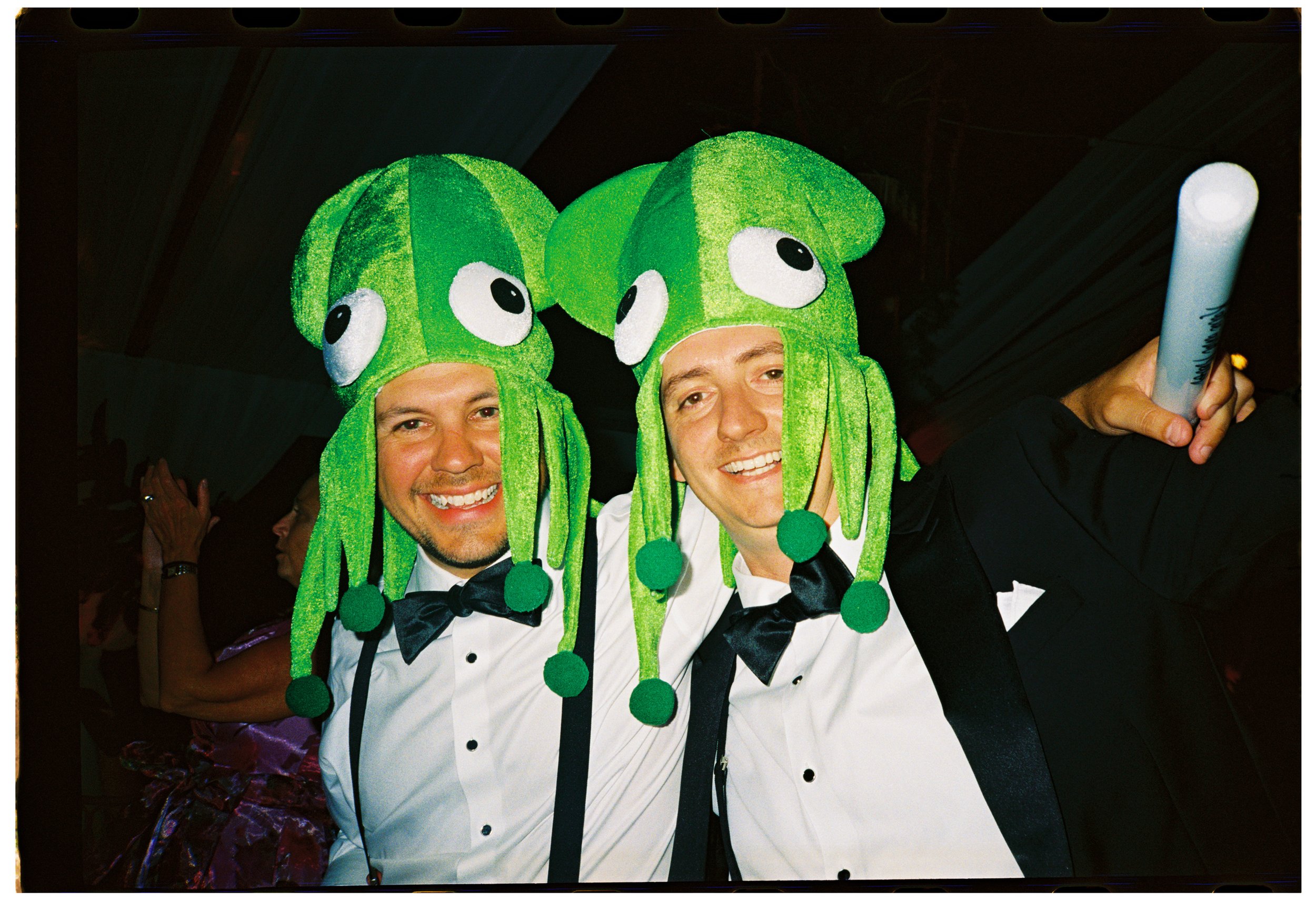 Black-tie wedding guest smiles in a playful green character hat during the late-night reception.