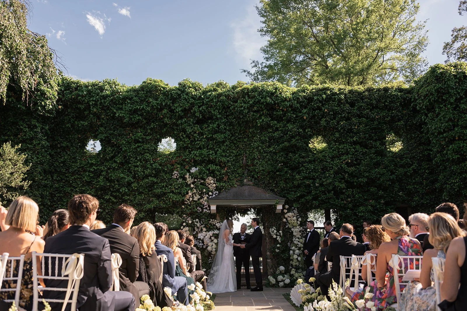 Outdoor wedding ceremony at the Ivy Wall at Goodstone Inn with guests seated beneath greenery and white floral installations.
